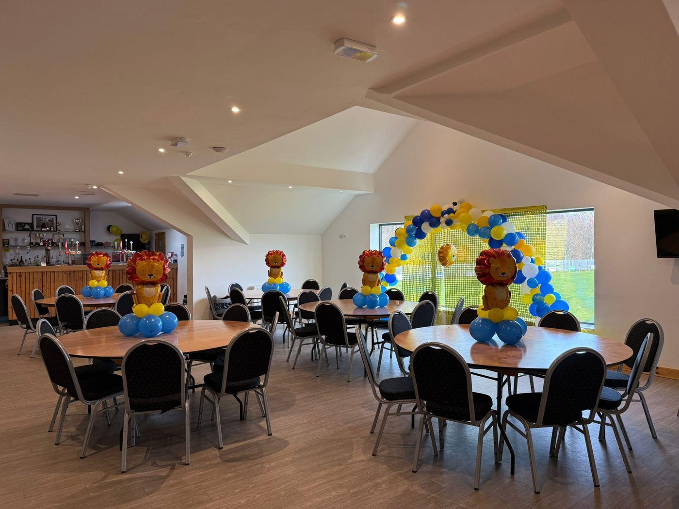 The clubhouse function room set up for a children's party, with round tables, lion balloon centrepieces and a balloon arch in front of a window over the pitch. The bar is at the back.