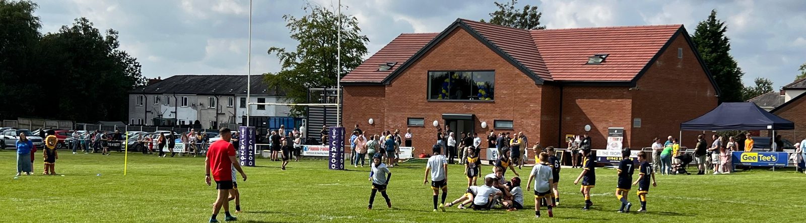 A junior match in progress on the pitch at The Den, with the goalposts and the clubhouse behind and supporters watching from the touchline.