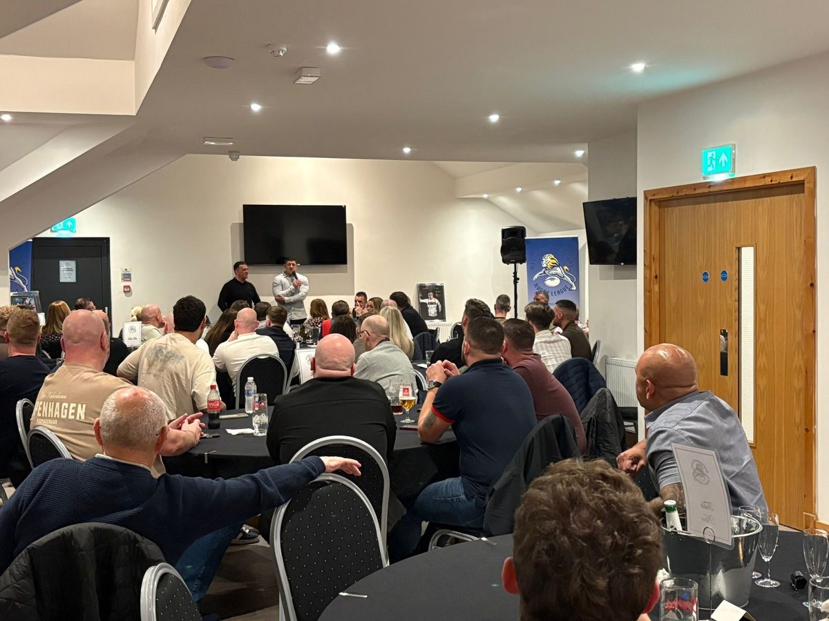 A speaker addressing a seated audience at the front of the function room, with wall-mounted screens and a PA system