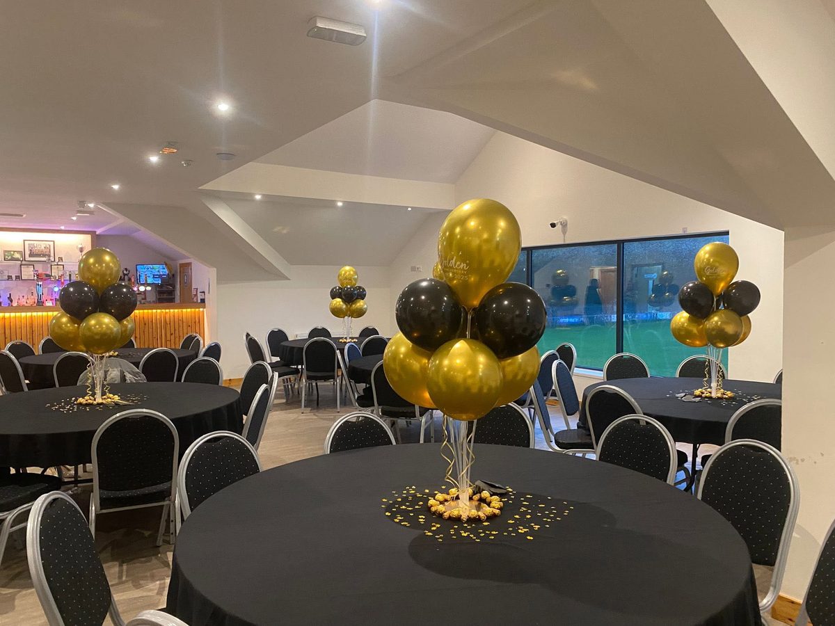 The function room laid out with round tables, each with a gold and black balloon centrepiece, ready for a celebration
